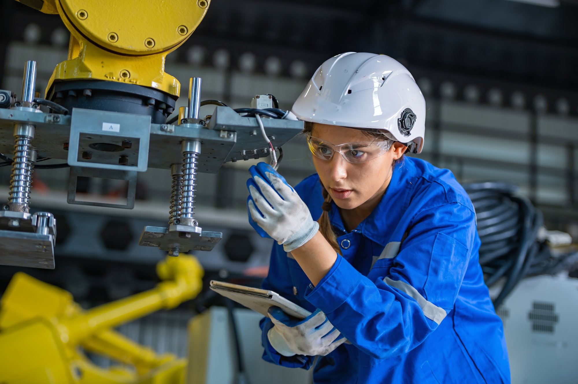 A female engineering technician in hard hat and safety goggles holds a notebook in one gloved hand while examining a machine with the other. 