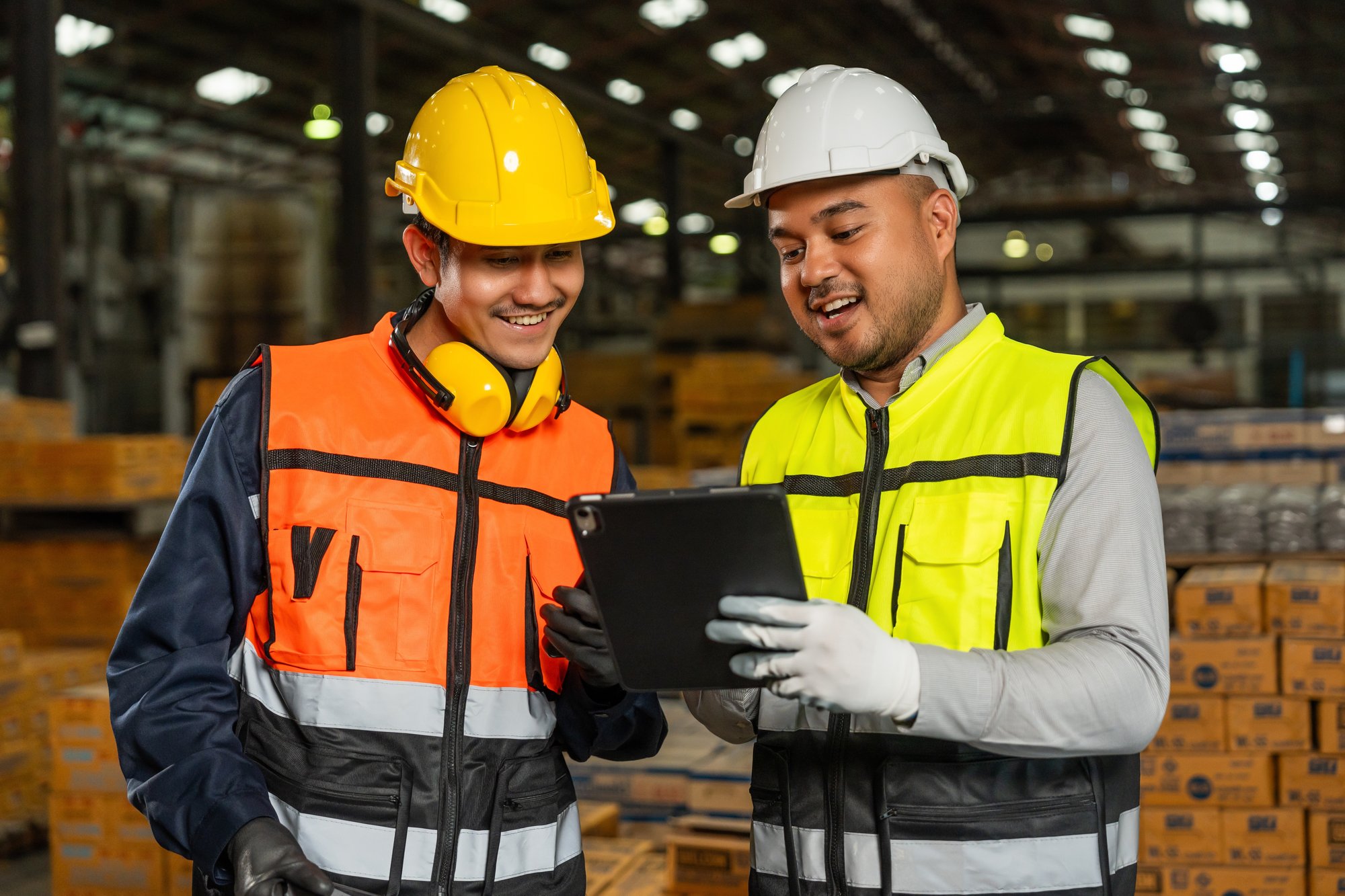 Two men in hard hats and bright safety vests stand in a warehouse looking at a tablet the man on the right is holding.